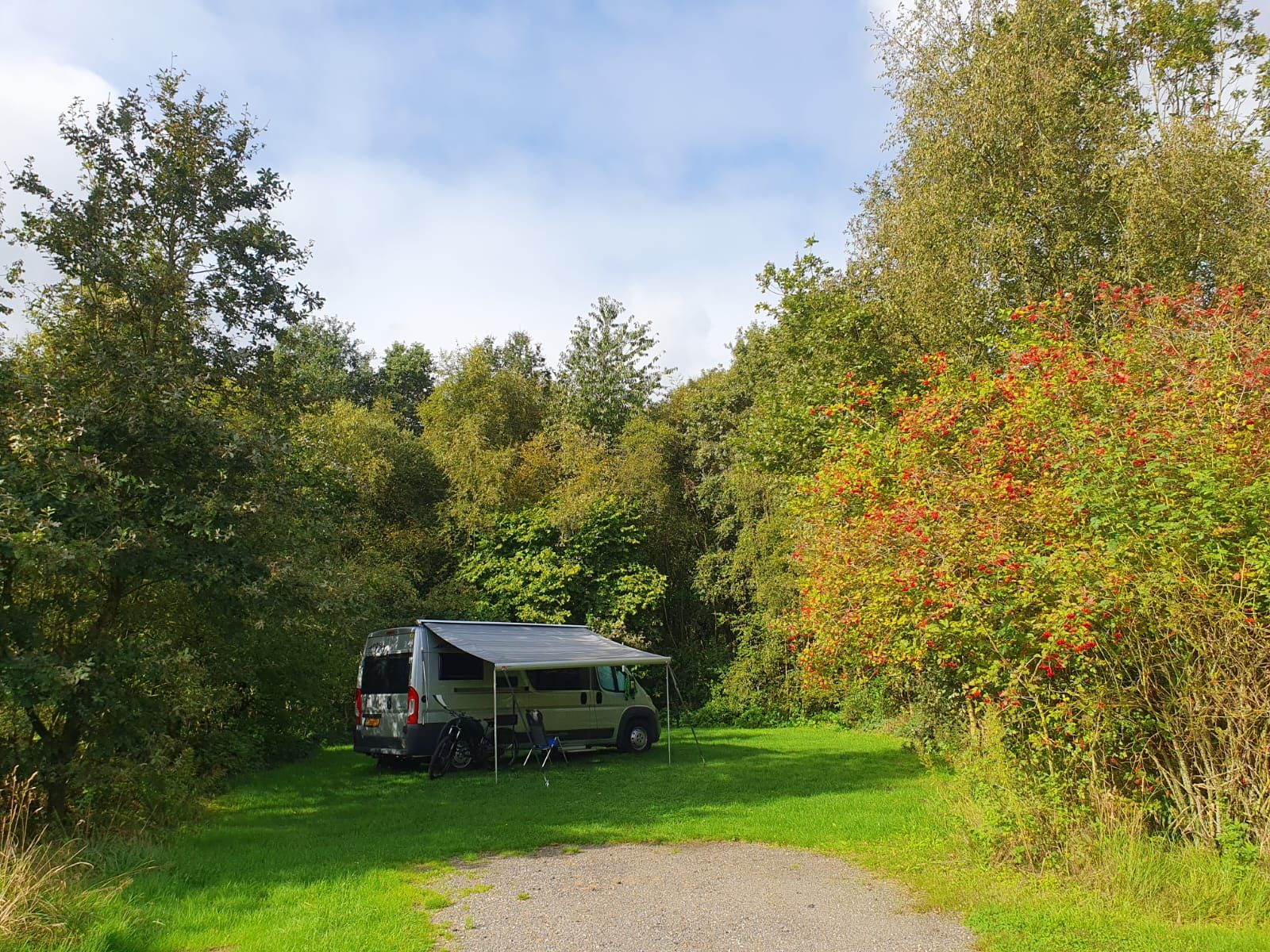 Camper met luifel op een ruime grasplaats aan een pad, tussen bomen en herfstkleurige struiken