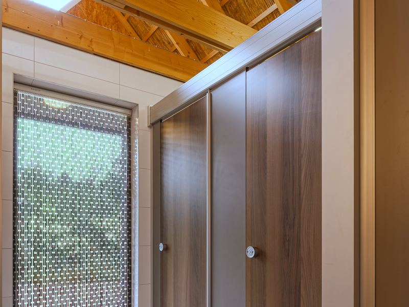Toiletruimte in het sanitairgebouw, met cabinedeuren in houtlook onder een open kap van houten spanten en riet